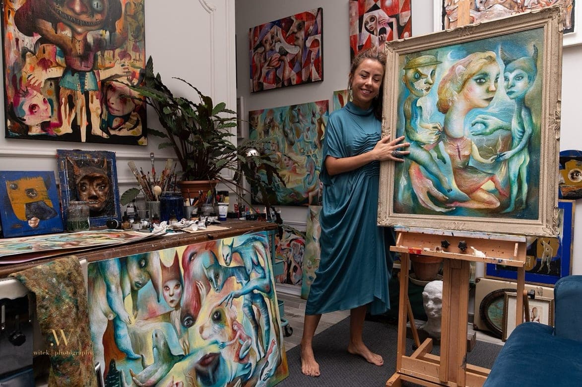 Valentina Brostean en su estudio, rodeada de sus pinturas, sosteniendo The Conversation Between Past, Present, and Future sobre el caballete